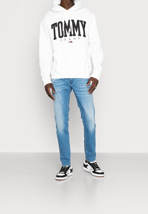Sweat à capuche blanc avec le texte "TOMMY JEANS" en noir, jean en denim bleu et baskets noires et blanches, debout devant un arrière-plan neutre.