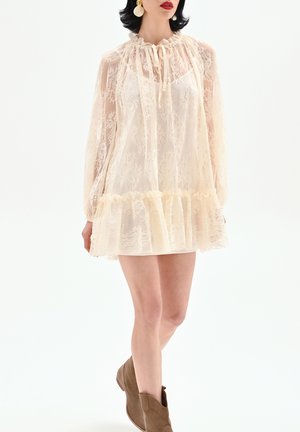 Femme portant une mini robe en dentelle crème à manches longues, ourlet volanté et col à nouer, associée à des bottines marron et des boucles d'oreilles rondes.