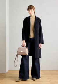 Czarny wełniany płaszcz, lekki sweter o fakturze, ciemne rozszerzane dżinsy, beżowa torebka na pasku i metaliczne buty na drewnianej podłodze.