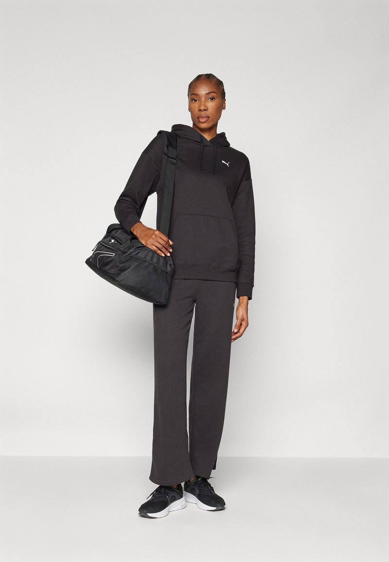 Schwarzer Kapuzenpullover und weit geschnittene Hose aus weichem Material. Trägt eine schwarze Tasche mit Netzakzenten und einem Schultergurt. Trägt schwarze Turnschuhe.