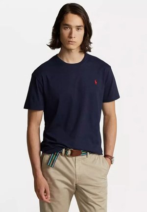 T-shirt in cotone blu navy con collo rotondo, maniche corte e un piccolo logo rosso ricamato sul petto; indossato con pantaloni beige e una cintura a righe.