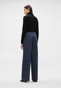 Sieviete stāv sāniski, ģērbta melnā augstā apkaklī un vaļīgās zilās biksēs, vienkārša gaiši pelēka fona priekšā.