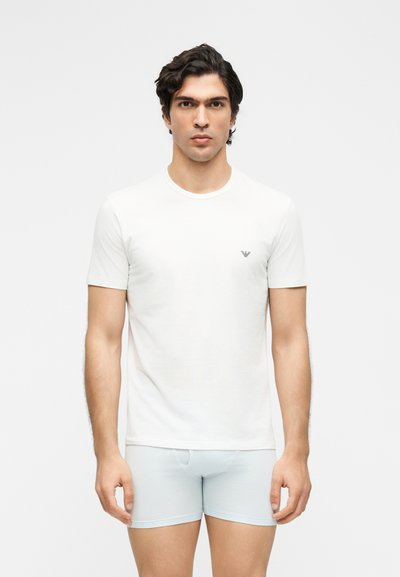 Άνδρας με σκούρα μαλλιά, που φοράει απλό λευκό κοντομάνικο μπλουζάκι και ανοιχτό μπλε μποξεράκια, στέκεται μπροστά σε λευκό φόντο.