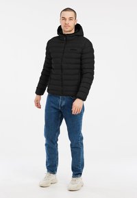 Zwart pufferjack met een capuchon, ritssluiting en geribbelde manchetten. Gecombineerd met blauwe spijkerbroek en crèmekleurige sneakers, staand op een witte achtergrond.