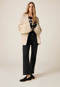 Beige chunky strikket cardigan med oversize ærmer, kombineret med sorte, vidde jeans og sorte glitrende loafers. Glat stof og afslappet pasform.