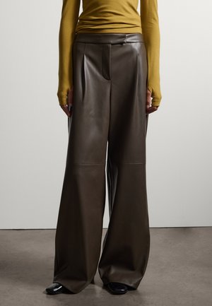 Persona che indossa pantaloni di pelle marrone scuro a gamba larga e una maglia a maniche lunghe color senape, in piedi su un pavimento grigio davanti a una parete semplice.