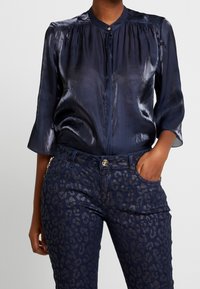 Blouse en satin bleu marine avec un col montant et des manches trois-quarts, associée à un pantalon en denim foncé avec un motif léopard subtil.
