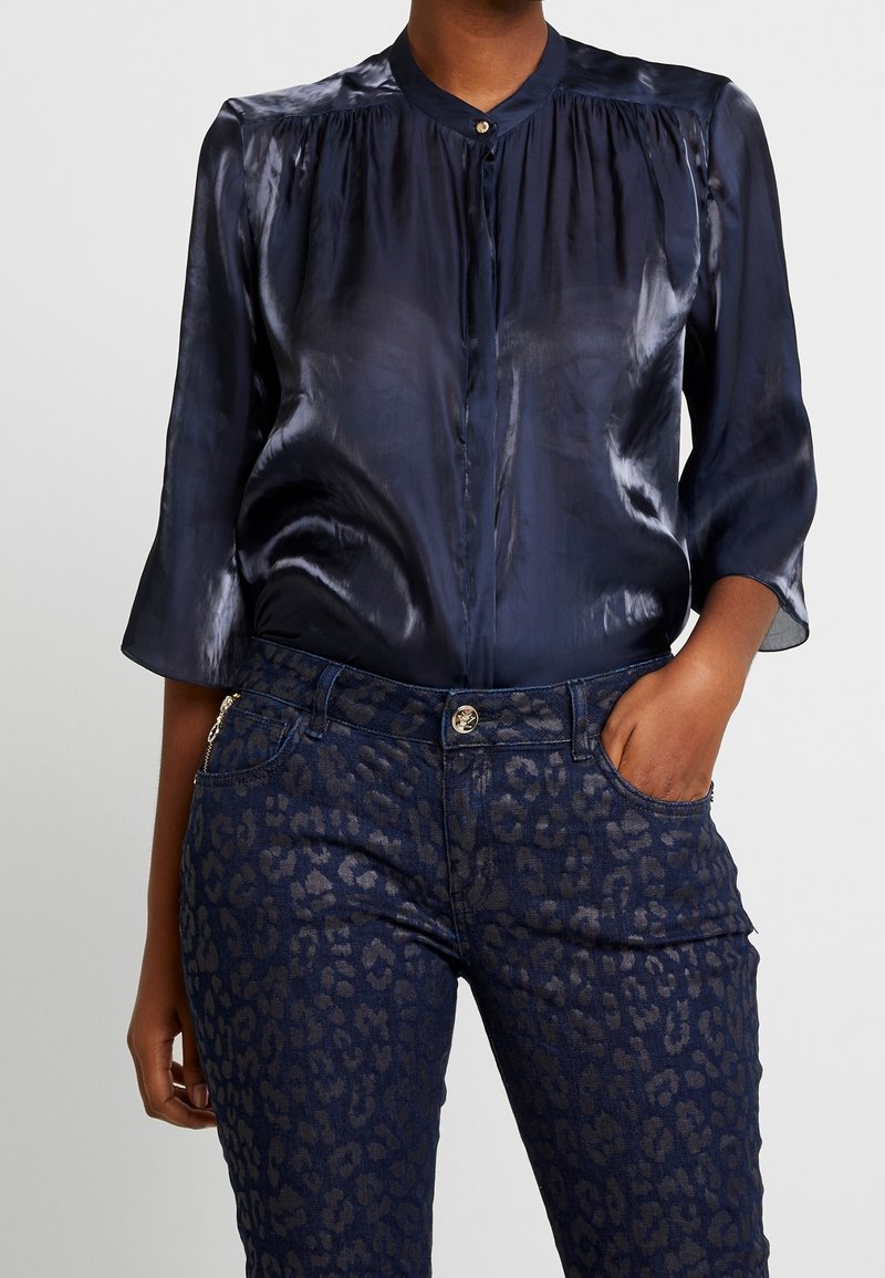 Blouse en satin bleu marine avec un col montant et des manches trois-quarts, associée à un pantalon en denim foncé avec un motif léopard subtil.