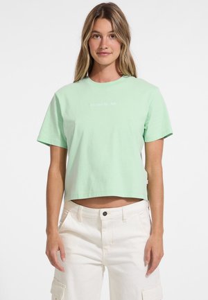 Mėtų žalia trumpa medvilninė marškinėliai, turi trumpas rankoves ir apvalų iškirpimą, su balta "GUESS. JEANS" logotipu.