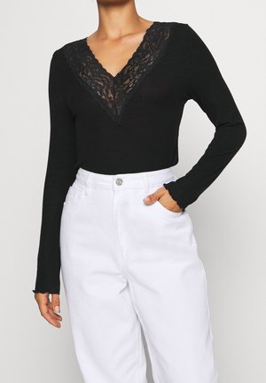 Femme vêtue d'un haut noir à manches longues avec col en V en dentelle et d'un pantalon blanc taille haute, une main dans la poche, sur fond uni.
