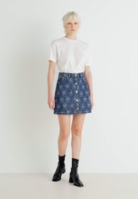 Denim mini krilo z modrim cvetličnim vzorcem, zlatimi gumbi spredaj, v skladu z enostavno belo majico in črnimi gležnjarji.