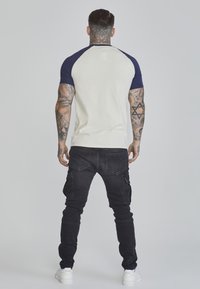 Kurzärmliges Raglan-T-Shirt in hellem Creme mit marineblauen Ärmeln. Verfügt über einen runden Halsausschnitt und wird mit schwarzen Cargo-Hosen und weißen Turnschuhen kombiniert.