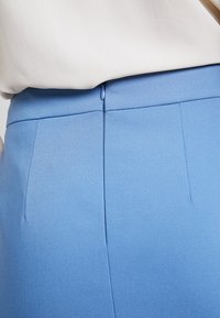 Pantalon bleu ajusté avec une texture lisse, comprenant une couture arrière et deux poches verticales. Le tissu semble doux et bien coupé.