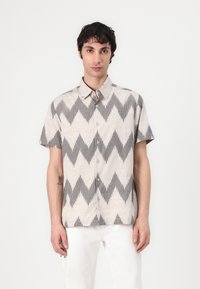Camicia a maniche corte con bottoni di colore beige con motivo a zigzag nero. Presenta un colletto a punta e un tessuto leggero, adatta per un abbigliamento casual.