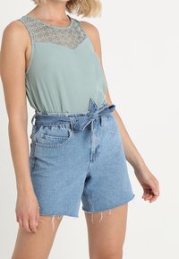 Ljusgrön ärmlös topp med spetsdetalj vid halsen, matchad med högmidjade blå denimshorts med bälte i midjan och fransad nederkant.