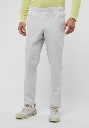Un pantalon de survêtement gris clair avec une taille élastique, une texture lisse et des poches latérales. Associé à des chaussures de sport grises et jaunes.