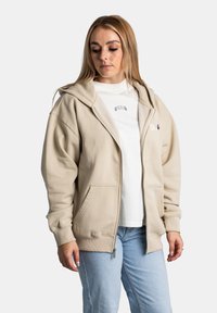 Sudadera con cremallera beige de textura suave, grandes bolsillos frontales, capucha con cordón y puños acanalados, superpuesta sobre una camiseta blanca.