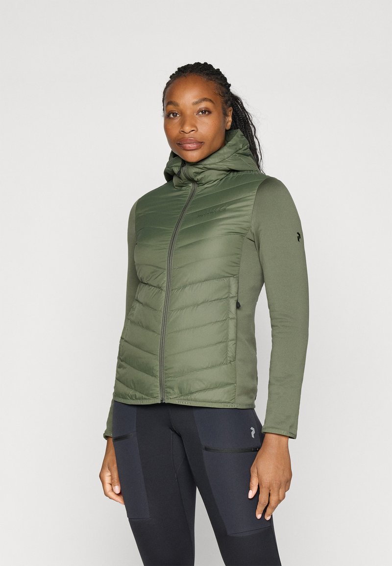 Giacca a vento verde con chiusura a zip e cappuccio. Il corpo presenta una texture trapuntata; le maniche sono lisce e aderenti. Sotto indossa pantaloni sportivi neri.