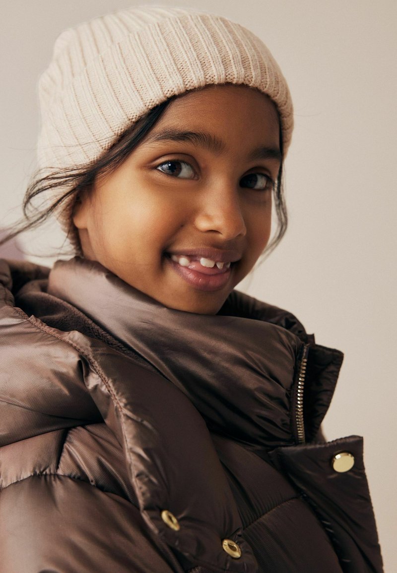 Braune Pufferjacke mit hohem Kragen und goldenen Details. Weiche beige Rippstrickmütze. Jugendlicher mit dunklem Haar und warmem Hautton.