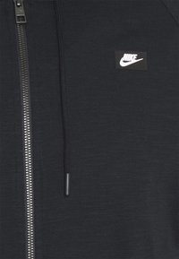 Svart zip-up hoodie med texturerad tyg. Har en silverfärgad dragkedja och en liten vit Nike-logotyp på en svart lapp. Inkluderar snören.