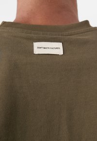 Olivengrøn bomulds-t-shirt med rund hals; mærket på bagsiden læser "DON'T WASTE CULTURE®" i et hvidt rektangulært mærke.