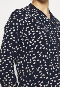 Kvinna iförd marinblå zip-up jacka med små vita och gula blommönster, delvis synlig ljusbrun skjorta under.