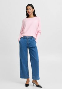 Pull en tricot rose clair, large pantalon en denim bleu avec ourlet effiloché, et escarpins noirs à bout pointu. Le mannequin se tient avec les mains dans les poches.
