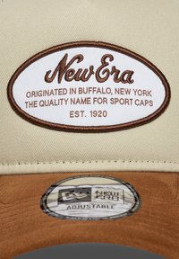 Gorra ajustable beige con un parche de logo cosido en marrón que dice "New Era". Parte inferior de fieltro suave y etiqueta circular con especificaciones.