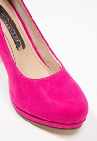 Gros plan sur une chaussure à talon haut en suède rose vif, avec un bout arrondi et une doublure intérieure beige sur fond blanc.