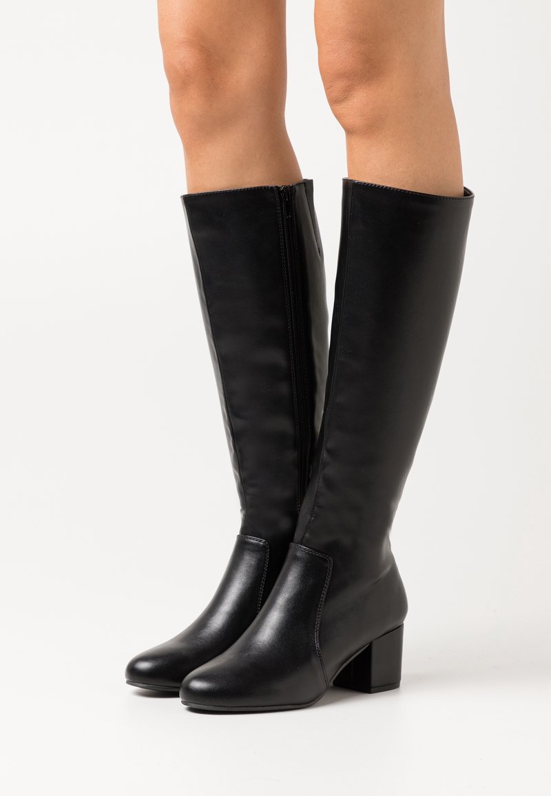 Bottes en cuir noires montantes jusqu'aux genoux avec talons blocs portées sur des jambes nues sur un fond clair uni.