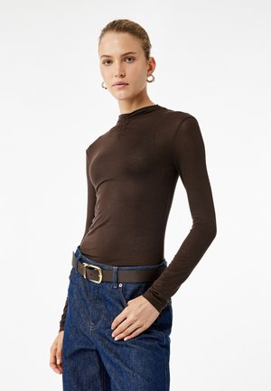 Bruine langarmpul met hoge hals en subtiele stikdetails, gedragen met blauwe spijkerbroeken en een bruine riem met een gouden gesp.