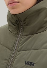 Chaqueta acolchada verde oliva con cuello alto, cierre de cremallera y textura acolchada. Presenta el logo negro de Vans en la parte frontal. Acabado de tela suave.