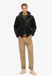 Sort og grå fleecejakke med hætte, lynlåslomme på brystet, båret over en grøn sweater, parret med beige bukser og lyse sneakers.