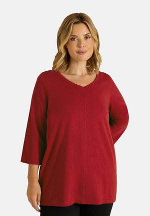 Vrouw met schouderlang blond haar, die een loszittend rood V-hals topje draagt met driekwart mouwen tegen een effen achtergrond.