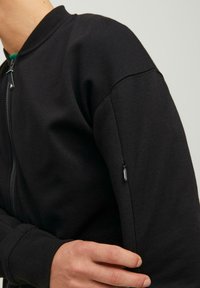 Črna bomber jakna na zadrgo iz teksturiranega materiala, z rebrastim ovratnikom in manšetami, ter majhnim žepom na zadrgo na rokavu.