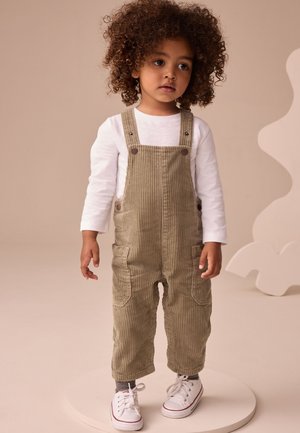 Bambino con capelli ricci che indossa una maglietta bianca a maniche lunghe, salopette beige in velluto a coste, calzini a righe e scarpe da ginnastica bianche, in piedi su una piattaforma rotonda.