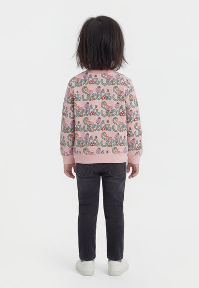 Copil stând cu fața spre spate, purtând un hanorac roz cu model floral colorat și text, pantaloni negri și pantofi albi pe fundal alb.