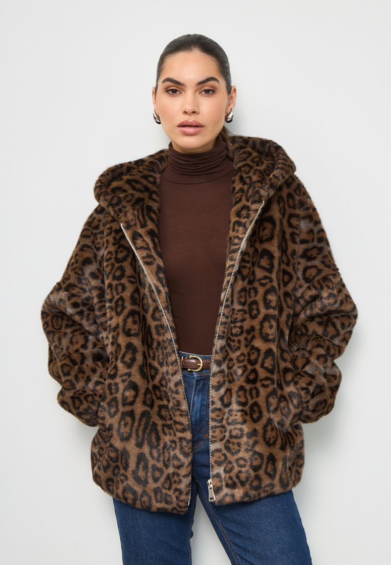 Manteau en fausse fourrure imprimé léopard marron avec fermeture éclair, coupe oversize et capuche. Porté par-dessus un col roulé marron, associé à un jean bleu.