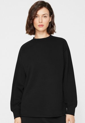 Femme aux cheveux courts et bruns portant un t-shirt noir uni à manches longues, debout sur un fond blanc.