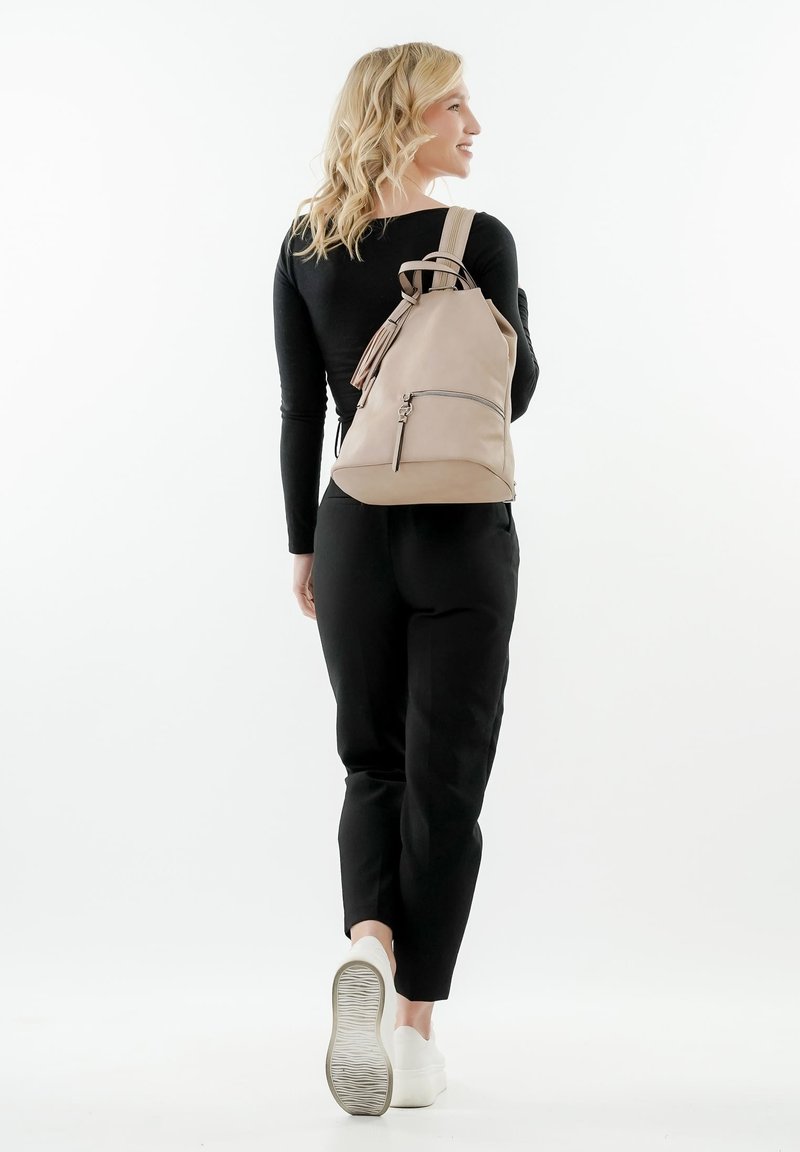 Zaino in pelle beige con spalline regolabili, una tasca frontale con zip e una base arrotondata, indossato da una persona in pantaloni neri e scarpe bianche.