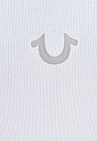 Un tessuto bianco a coste presenta un accento in rete grigia con un logo che somiglia a una 'U' cucito sul materiale. La texture è liscia e discreta.