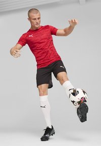 Camiseta atlética de manga corta roja con patrón texturizado, pantalones cortos negros, calcetines blancos hasta la rodilla y botas de fútbol negras, haciendo malabares con un balón de fútbol multicolor.