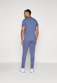 Blaues T-Shirt mit kurzen Ärmeln und Rundhalsausschnitt, mit dunkler Schrift auf dem linken Ärmel, kombiniert mit lila Sport-Jogginghosen und weißen Sneakers.