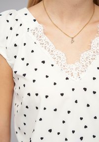 Weiße Bluse aus Stoff mit schwarzen Herzmustern, Spitzeneinfassung am Ausschnitt und kurzen Ärmeln. Besonderheiten sind ein subtil ausgefranster Rand.