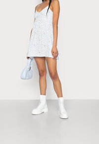 Blå blommig kortklänning med tunna axelband, bärs med vita kraftiga snörstövlar. Tillbehören inkluderar en ljusblå handväska. Slät tygtextur.