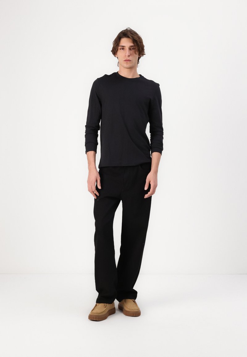 Sort langærmet skjorte parret med løse sorte bukser og beige slip-on sko, der viser et afslappet fit og minimalistisk design.