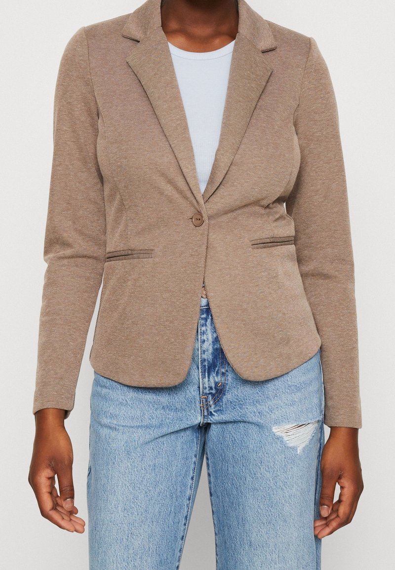 Femme portant un blazer ajusté marron clair sur un top blanc et un jean bleu clair déchiré, debout, les bras détendus le long du corps.