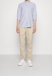 Camicia a righe blu e bianche con colletto, pantaloni khaki e sneakers bianche, in piedi su uno sfondo semplice.