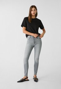 Sort kortærmet t-shirt, lysegrå slim-fit jeans og sorte spidse slingback-sko med dekorative hardware-detaljer.