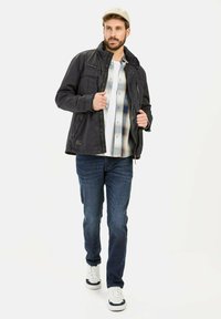 Veste noire avec des poches zippées, superposée à une chemise à carreaux. Associée à un jean foncé et des baskets blanches. Homme marchant sur un fond uni.
